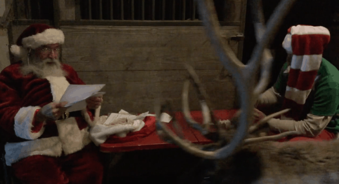 Santa reads a letter while reindeer walks in front of camera and Elf Wesley works on a Christmas ornament.