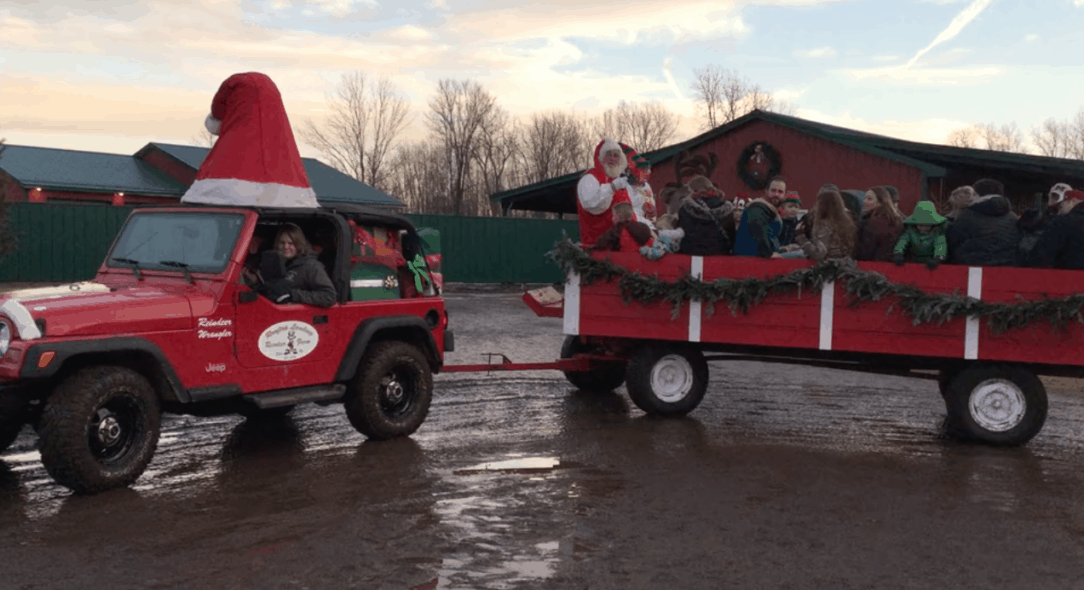 Red Jeep with large Santa hat on roof pulls Santa, Rudy, Skittles, and friends in a Christmas wagon. Sunset and Rooftop Landing barn in background.